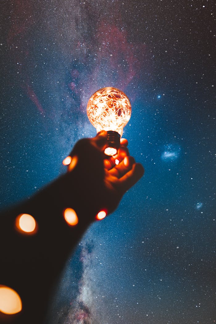 A hand holds a glowing bulb into the Milky Way night sky, creating a magical cosmic connection.